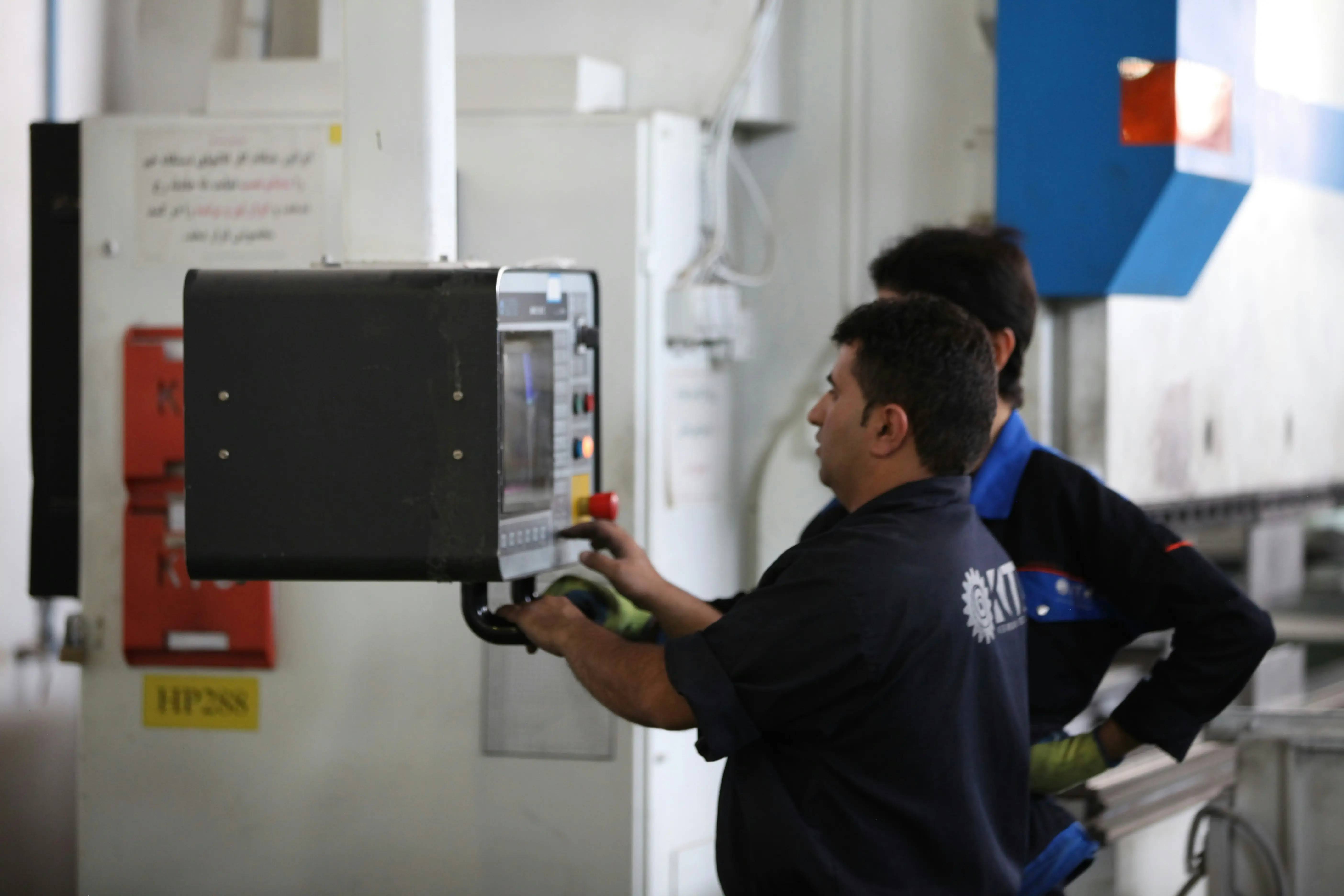 An operator working a machine control pendant on the shop floor, a colleague looking on